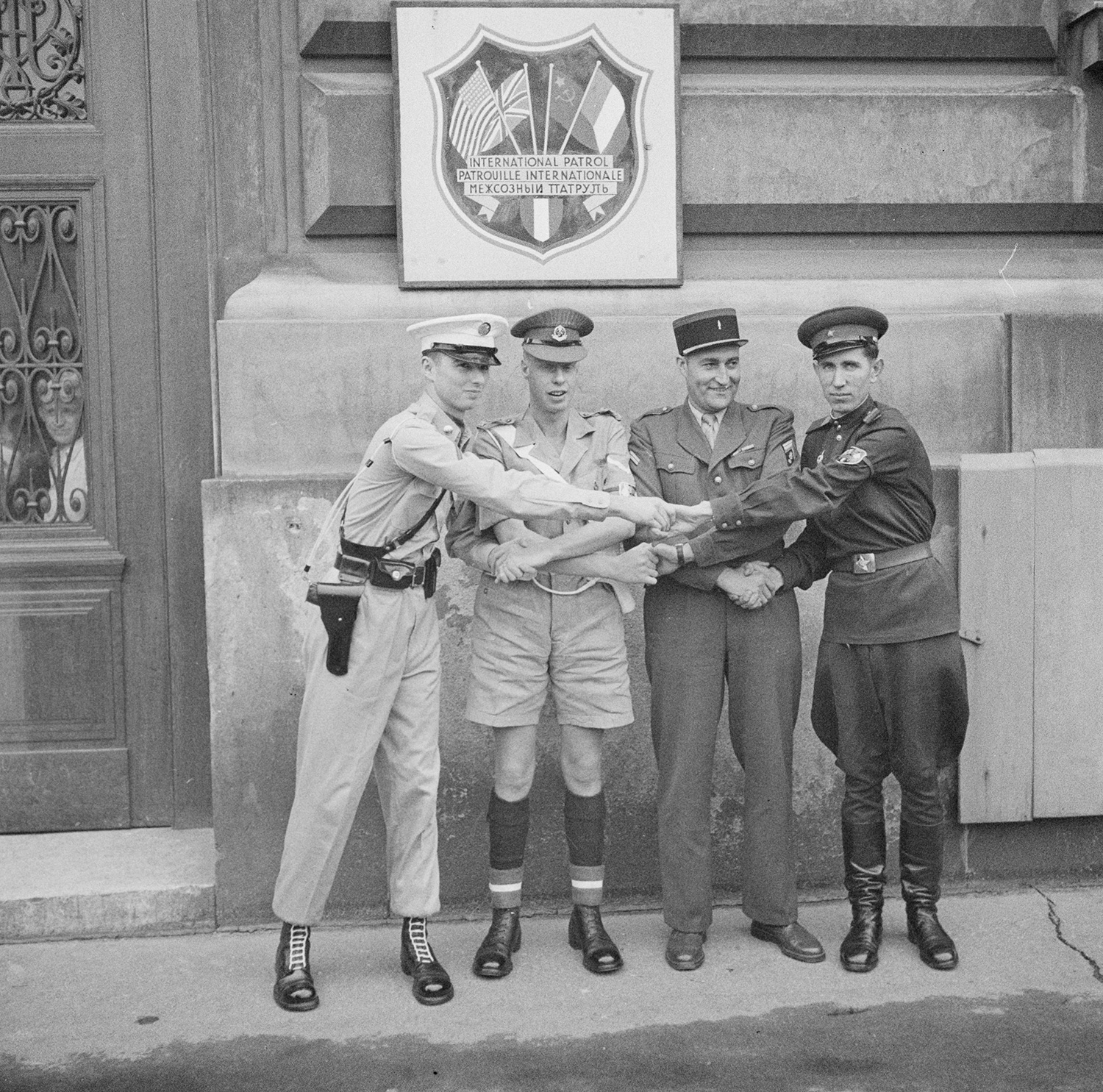 Schwarz-Weiß-Fotografie: Vier Soldaten der Alliierten in unterschiedlichen Uniformen geben sich vor einem Schild der International Patrol die Hände.