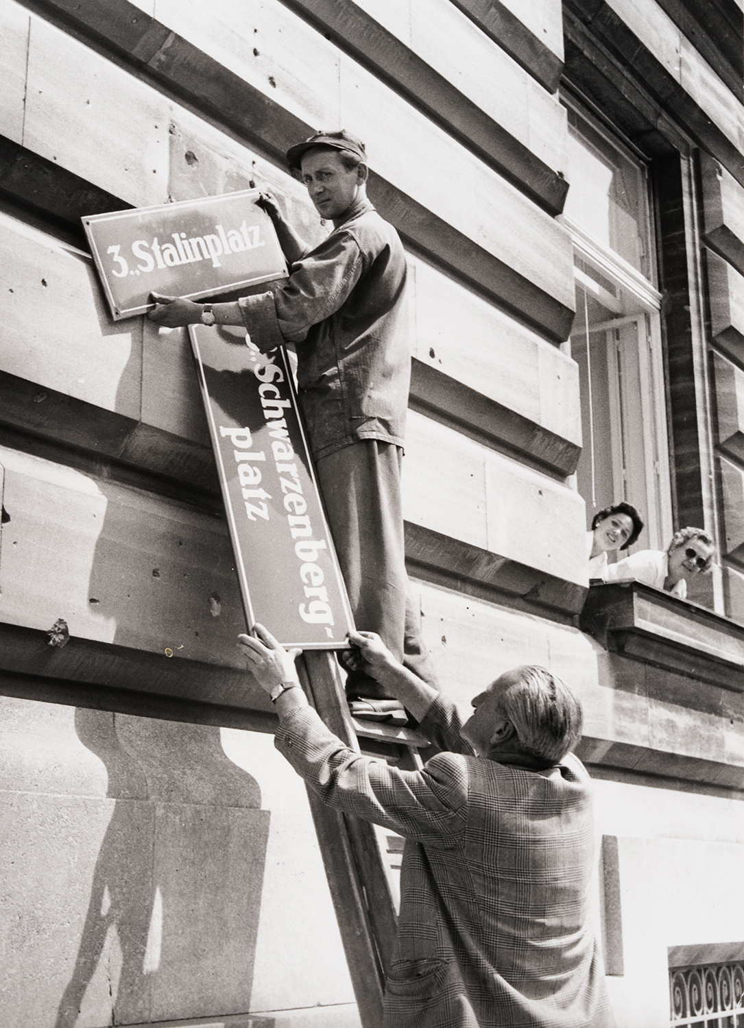 Schwarz-Weiß-Fotografie: Ein Mann hängt ein Straßenschild mit der Aufschrift „Stalinplatz“ ab, während ein anderer ihm das Schild „Schwarzenbergplatz“ reicht.