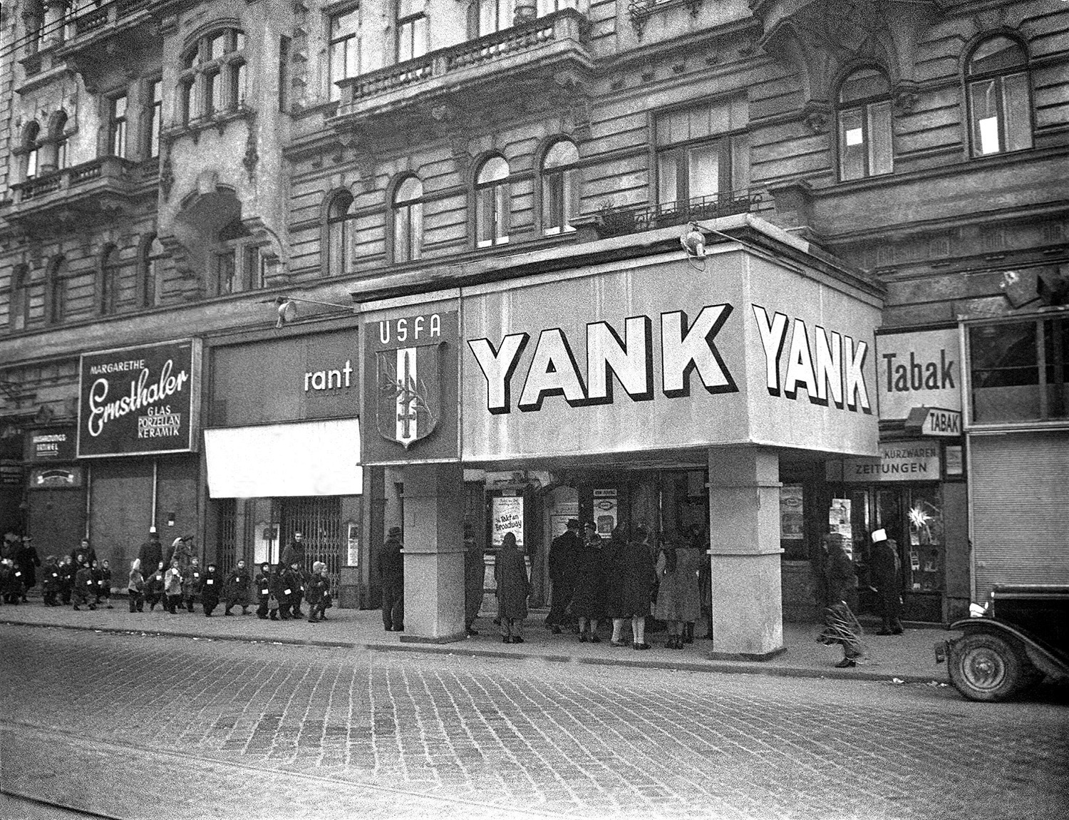 Schwarz-Weiß-Fotografie: Außenansicht des „Yank-Theatre“ in einem historischen Gebäude, mit Passanten und einer Gruppe Kinder auf dem Gehweg davor.