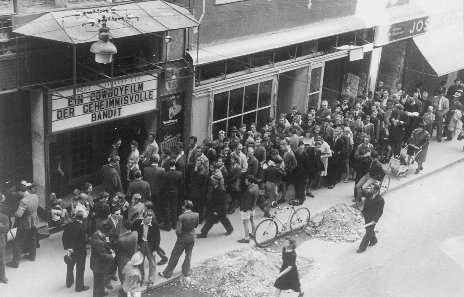 Schwarz-Weiß-Fotografie: Lange Menschenschlange vor einem Kinoeingang mit der Aufschrift „Ein Cowboyfilm: Der geheimnisvolle Bandit“ in belebter Straße.