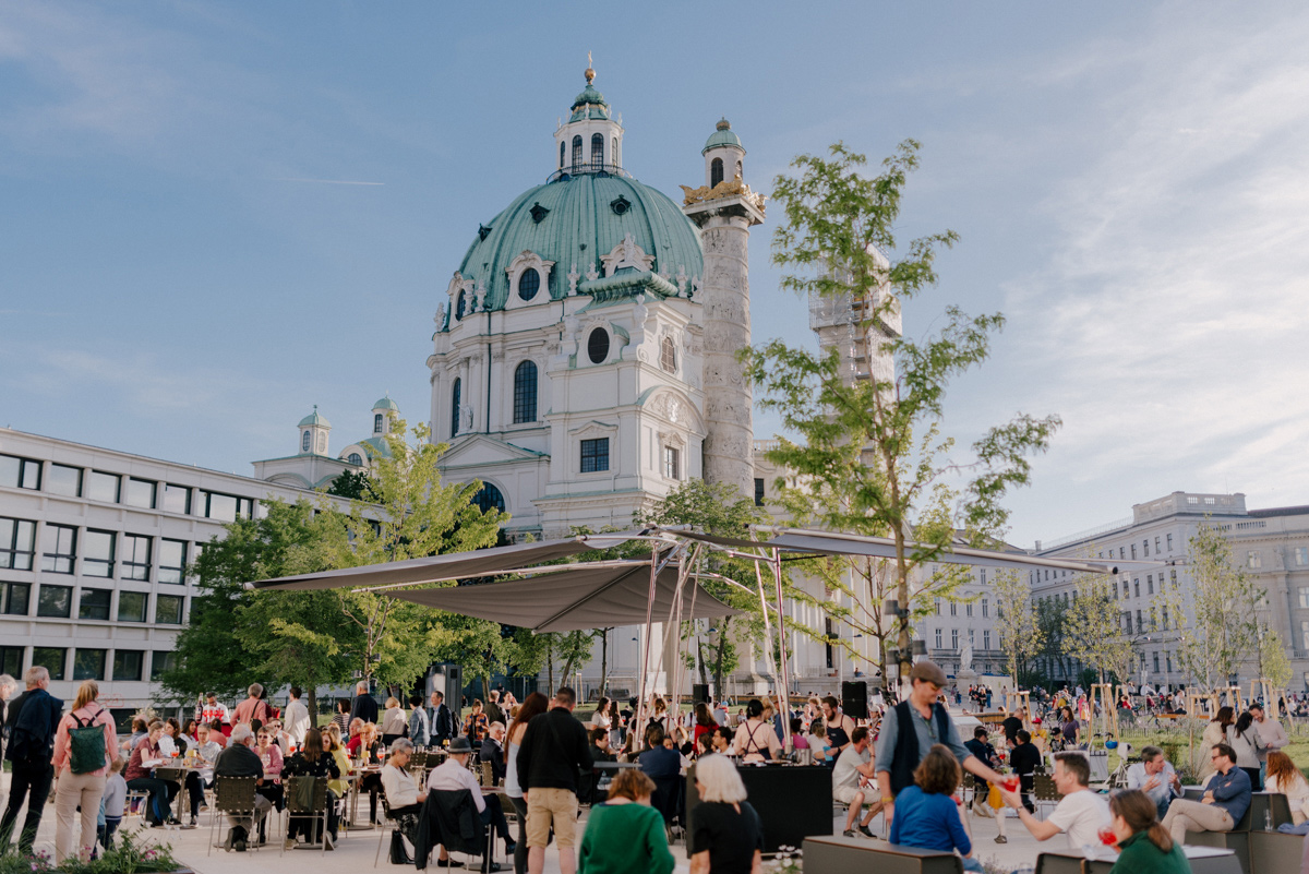 Gäste genießen Speisen und Getränke im Schanigarten des Wien Museums, im Hintergrund die Karlskirche